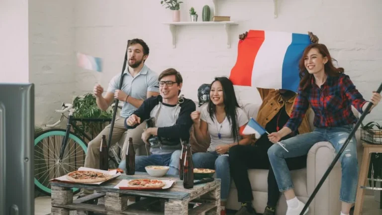 Group of friends cheering indoors with flags and pizza, enjoying a sports event.