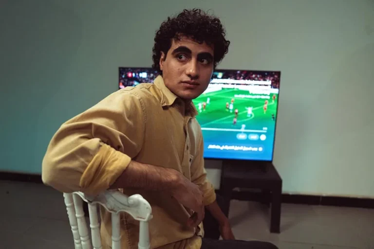 A young man sitting in a room, focused on a soccer match broadcast on TV.