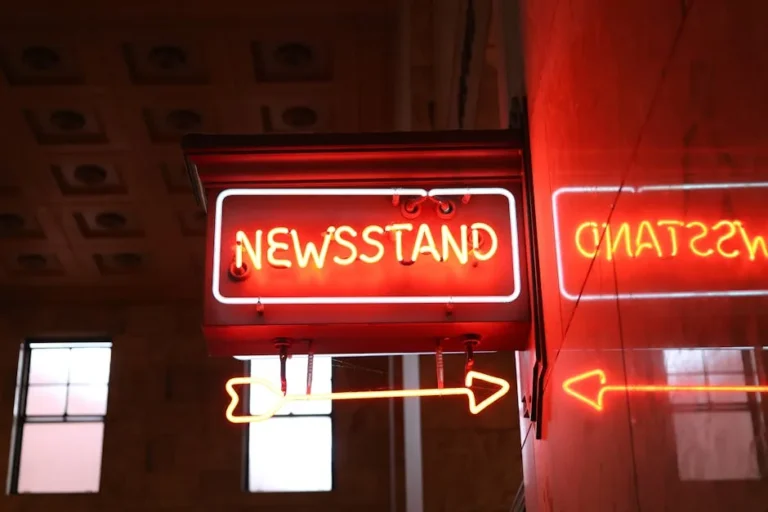 Bright neon newsstand sign reflecting on wall in urban environment.
