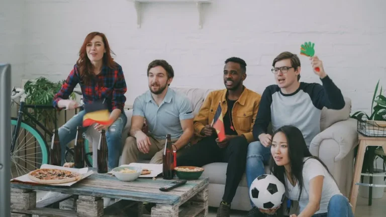 Friends watching a soccer match indoors, waving flags and cheering with snacks and drinks.