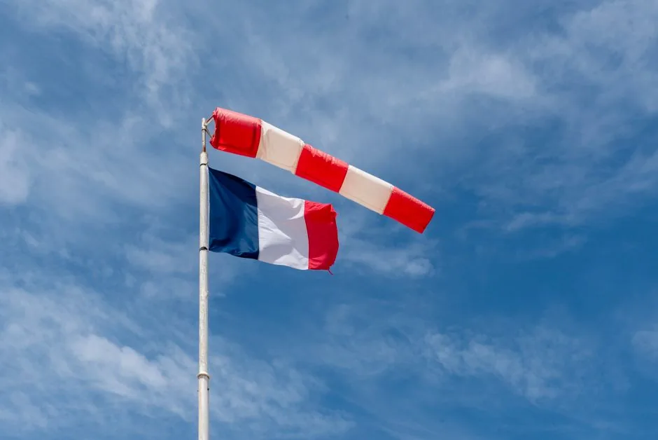 French flag and red-white windsock flying under a blue sky with scattered clouds.